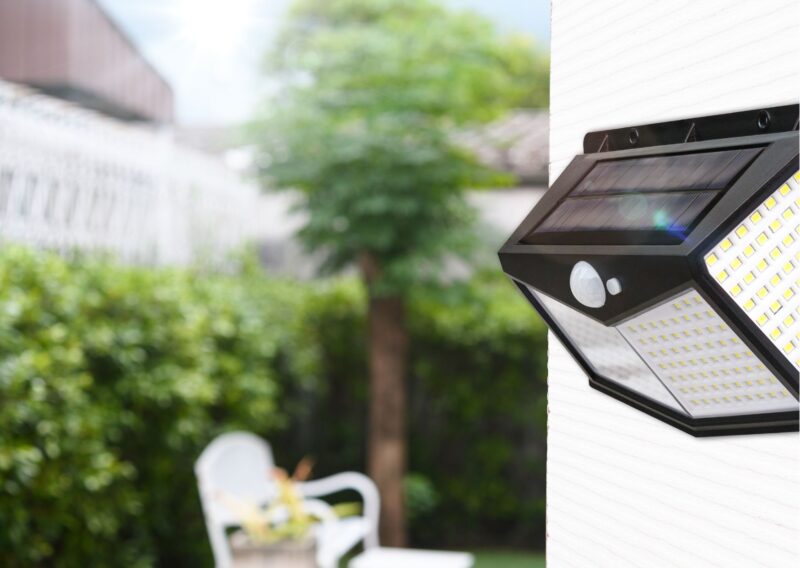 Lampada solare da esterno montata su una parete, dotata di pannello solare, sensore di movimento e luci LED, con giardino sfocato sullo sfondo